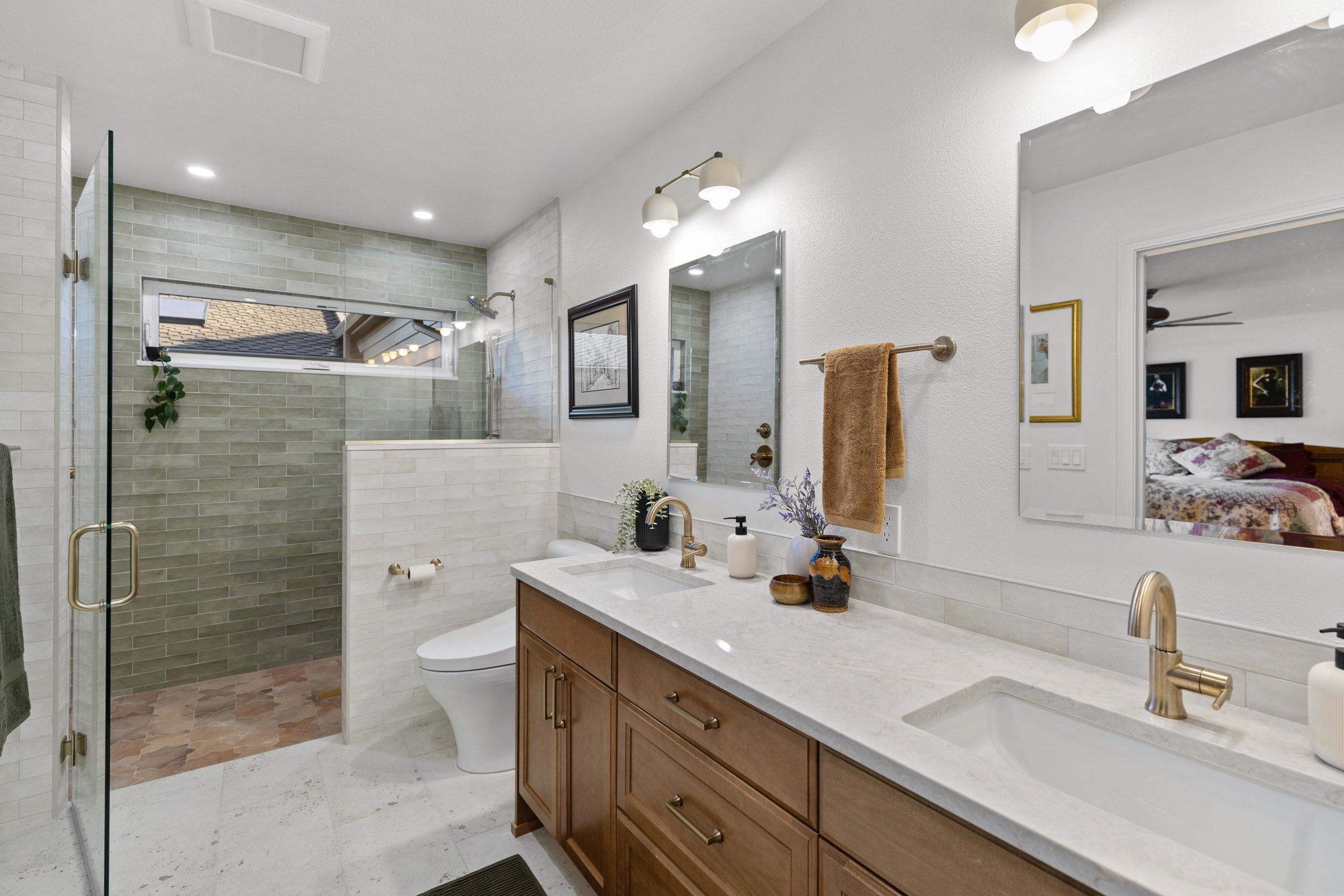 Accessible primary bathroom with a curbless walk-in shower in green tile and terracotta floor, a long window, and a double vanity with brass faucets