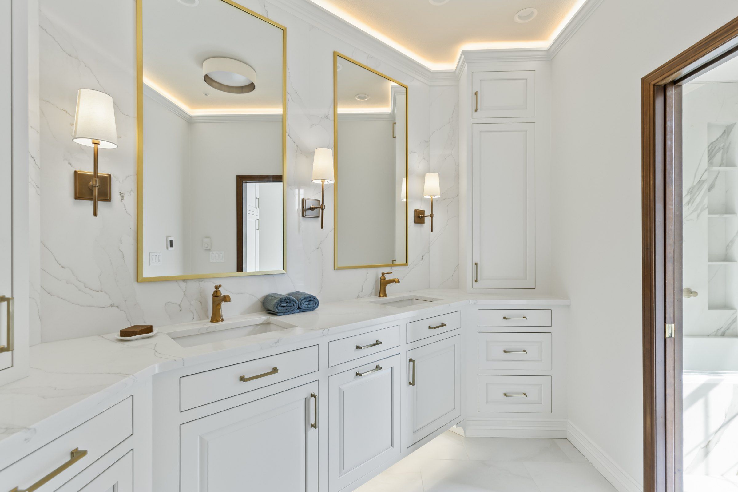 White bathroom with a double vanity, brass-framed mirrors, brass faucets and wall sconces
