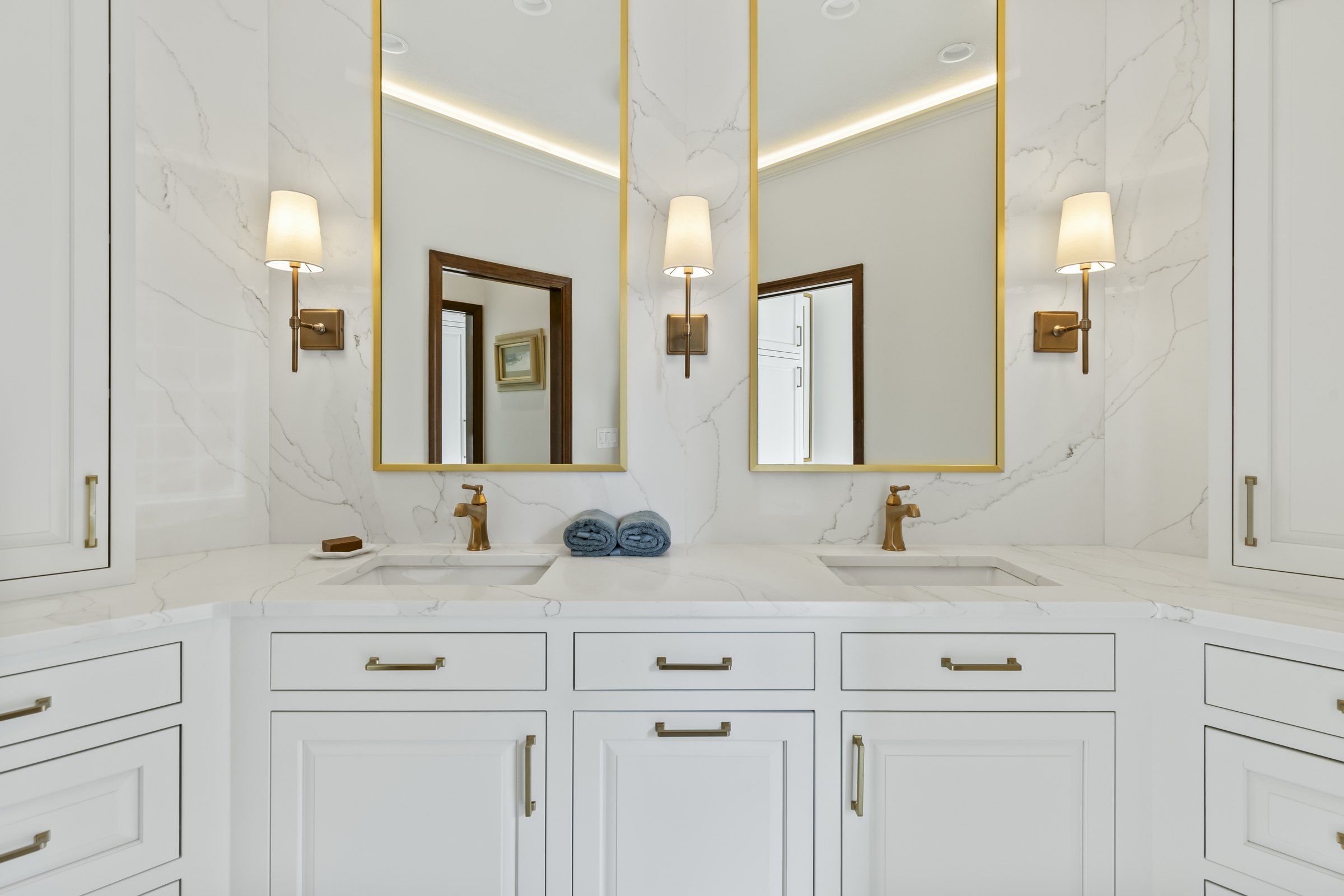Double vanity with brass mirrors and sconces under a lit cove ceiling