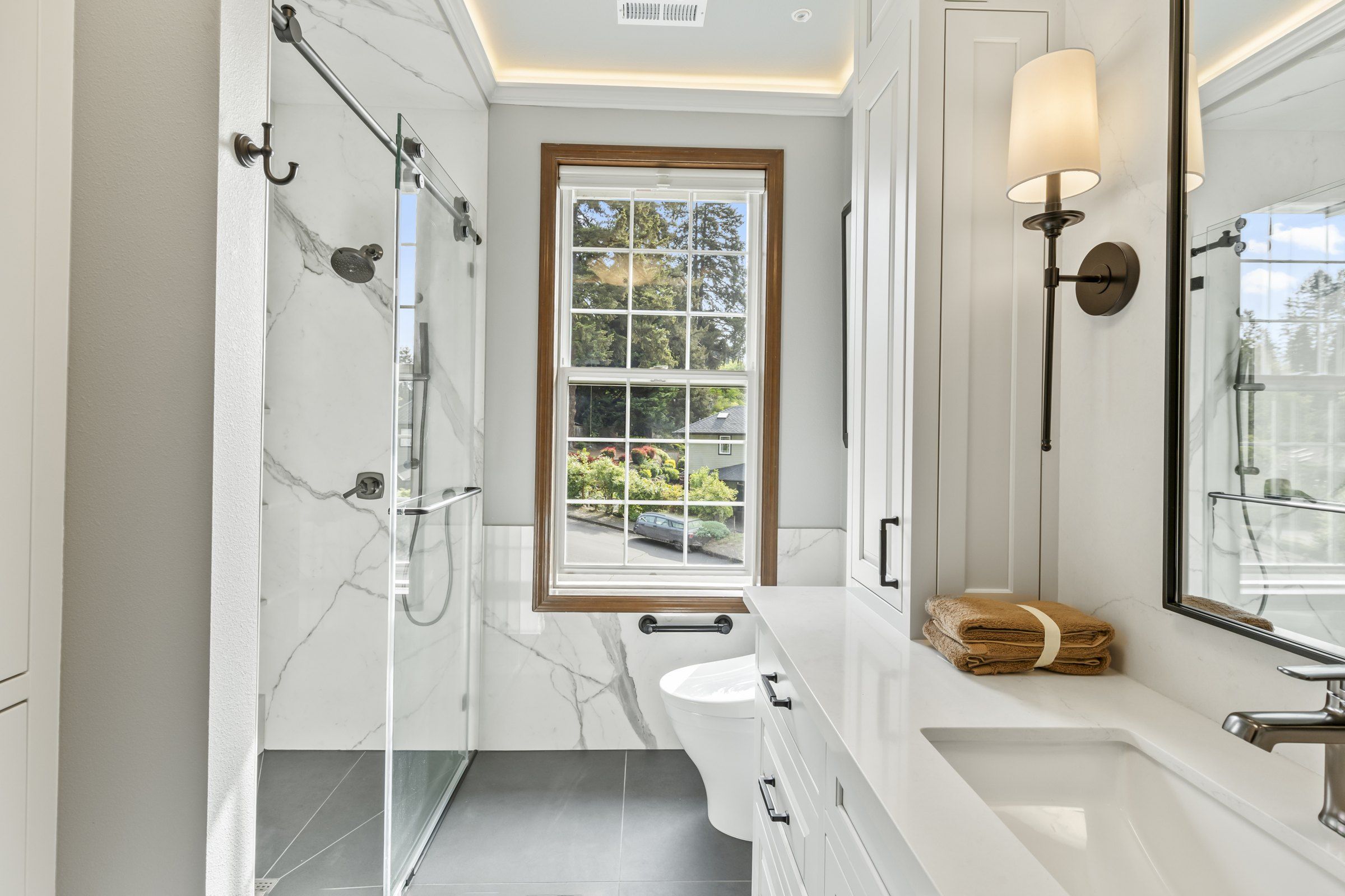 Glass-enclosed walk-in shower with marble-look wall panels beside a bidet toilet and window