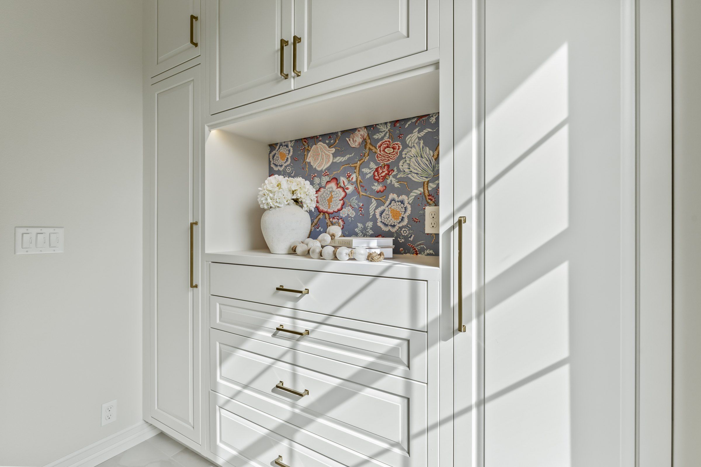 Walk-in linen closet with white cabinets, brass pulls and a wallpapered display niche